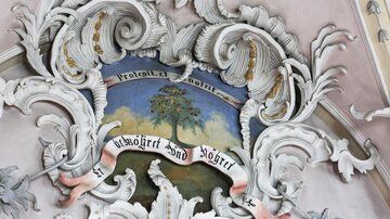 An artistic ceiling painting featuring a tree and curved embellishments. Below it is a banner with the text "Protest et munus".