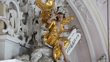 A golden statue of an angel sits on a cloud and holds a trumpet. The angel has outstretched wings and wears a historic garment.