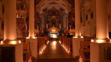 An atmospheric church with subdued light and ornate columns. The altar is visible in the background and illuminated by candles.