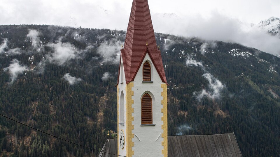 A church with a red roof and a tall bell tower in front of a mountainous landscape. The surroundings are characterized by green forests and gentle hills. | © Bernhard Gruber – Photography