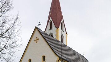 A beautiful church with a pointed red tower and a bright facade paint. In the foreground, there are some trees and a fence visible. | © Bernhard Gruber – Photography