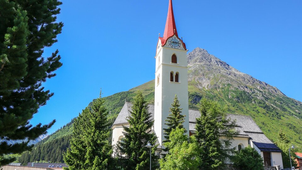 Eine malerische Kirche mit einem roten Turm, umgeben von grünen Bäumen und Bergen. Der Himmel ist klar und blau. | © TVB Paznaun - Ischgl