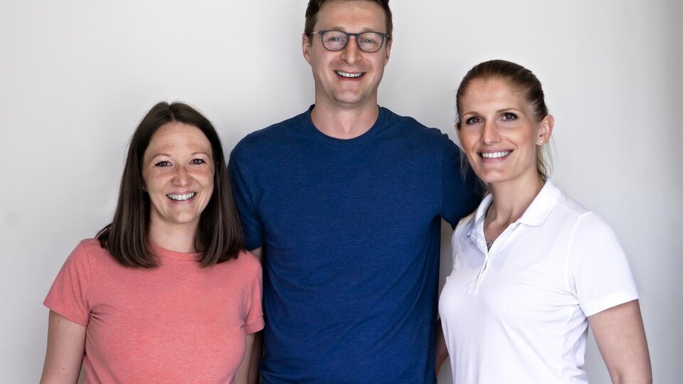 Three smiling individuals stand in front of a bright wall. They wear casual clothing and appear friendly and open.