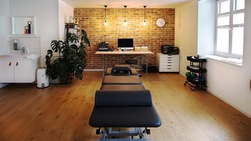 A modern practice room with a treatment table and stone wall. The room is brightly and invitingly designed, with a plant and workspace.