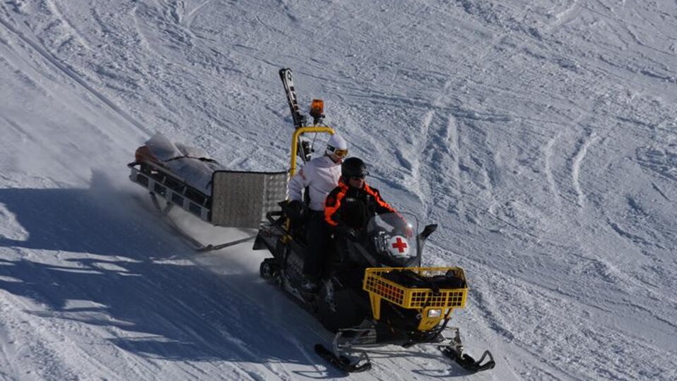 A snowmobile is driving over the snow. On the snowmobile, there are two people transporting equipment for medical emergencies.