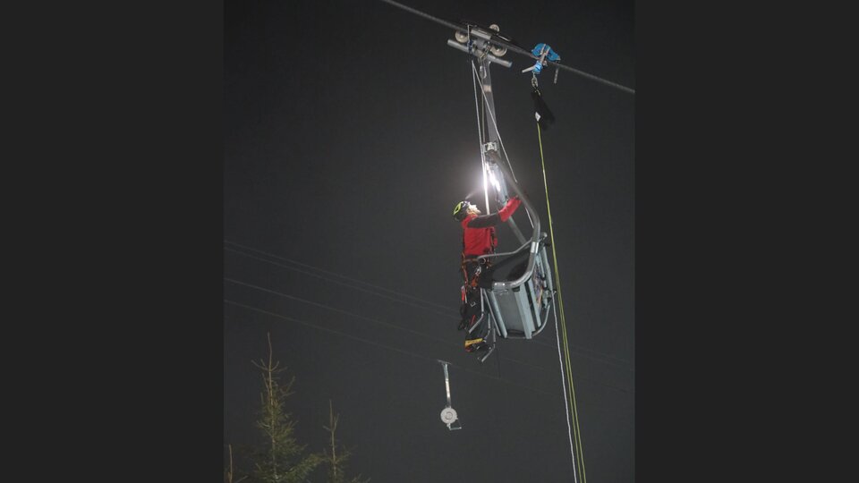 Eine Person arbeitet an einer Seilbahn in der Nacht. Sie trägt eine Sicherheitsausrüstung und nutzt eine Taschenlampe.