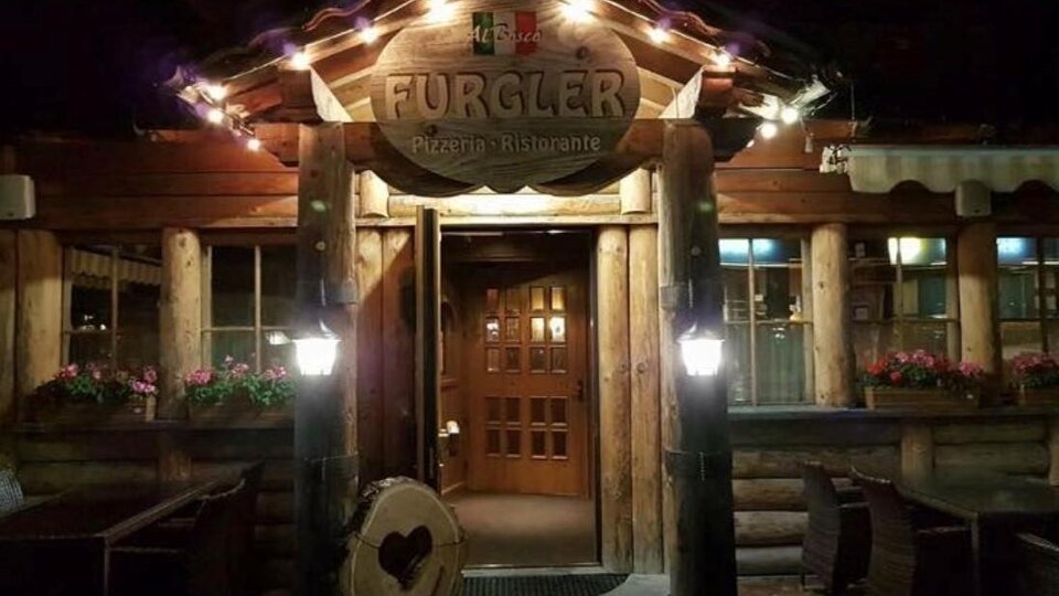 A rustic wooden restaurant with an inviting facade and warm lighting. The entrance door is made of wood and the windows are adorned with blooming plants.