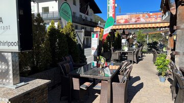 An inviting terrace of a restaurant with tables and chairs. In the background, green trees and a blue sky can be seen.