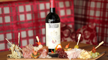 A bottle of red wine stands on a table, surrounded by a selection of sausage and cheese platters. The background features a cozy, rustic wooden paneling.