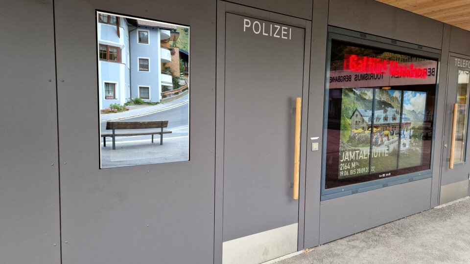 A modern police station entrance with a large window. Next to the entrance is a picture of a bank and residential buildings.
