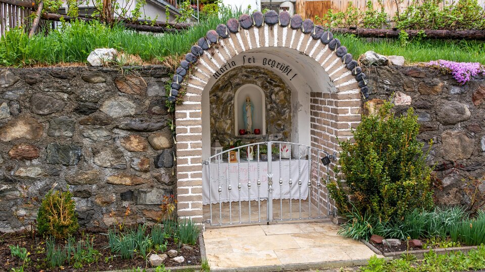 A small altar shrine in a stone wall with a white gate. Surrounded by green plants and flowers. | © Bernhard Gruber – Photography