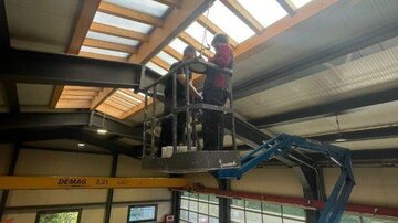 Two workers are standing on a lift and repairing the roof of a large industrial building. In the background, workshop machines and large windows can be seen.