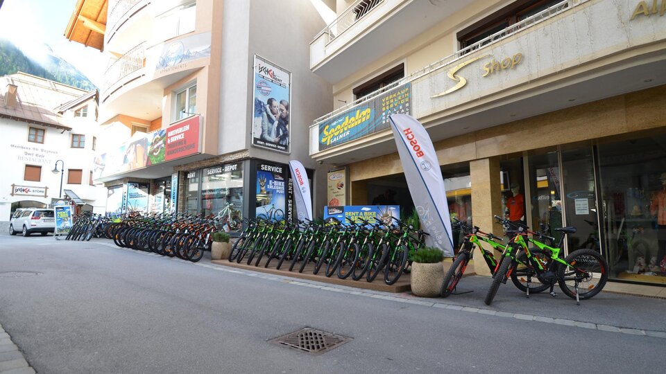 Eine Straße mit mehreren Fahrrädern, die zum Mieten bereitstehen. Im Hintergrund sind Shops und Werbefahnen sichtbar.