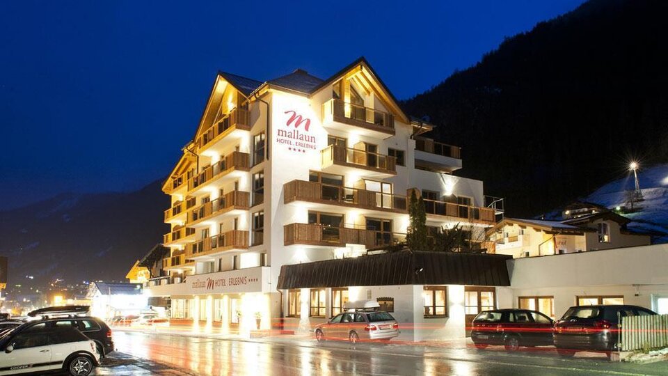 A modern hotel building with several stories and balconies. The surroundings are illuminated in the evening, and the streets are wet from the rain.