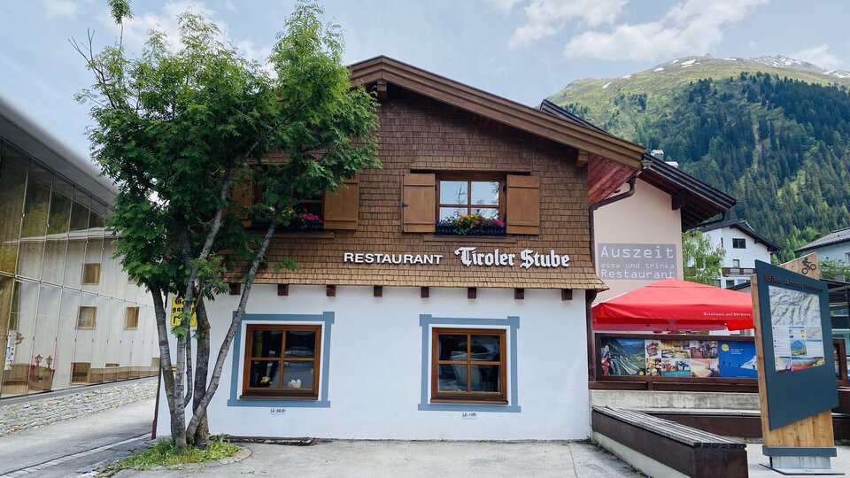 A cozy restaurant called "Tiroler Stube" with wooden paneling and windows. The mountains are visible in the background.