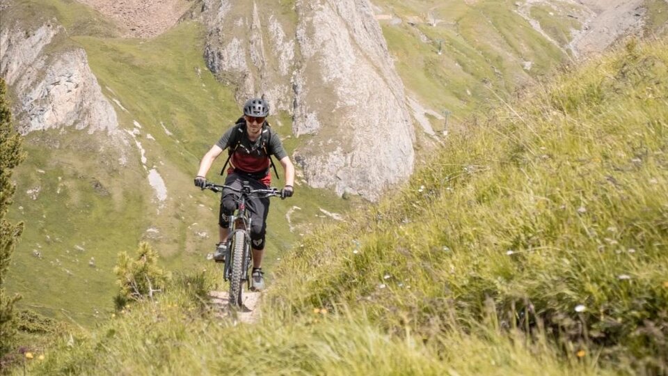 Ein Radfahrer fährt auf einem schmalen Weg durch die grüne Berglandschaft. Im Hintergrund sind felsige Berge und ein klarer Himmel zu sehen.