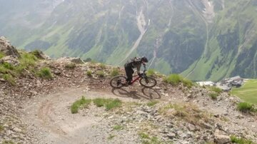 Ein Radfahrer fährt auf einem kurvigen Pfad in den Bergen. Im Hintergrund sind grüne Wiesen und majestätische Berggipfel zu sehen.