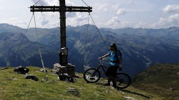 Eine Person steht mit ihrem Fahrrad an einem Gipfelkreuz in den Bergen. Im Hintergrund sind grüne Hügel und eine klare blaue Himmel sichtbar.