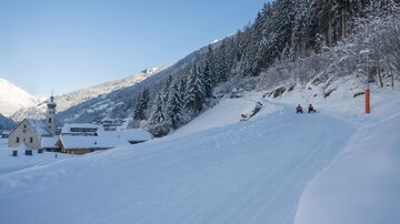 A winter landscape with snow-covered hills and fir trees. In the background, small houses and a church can be seen.
