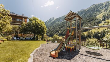 A playground with a slide and swings in a green meadow. In the background, mountains and a blue sky are visible.