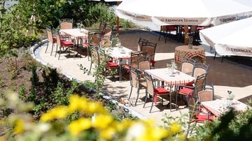 An inviting outdoor restaurant with tables and chairs under white parasols. The surroundings are green and blooming, perfect for relaxing and enjoying.