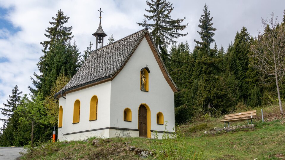 A small, white chapel stands amid trees. The sky is cloudy and the surroundings are green. | © Bernhard Gruber – Photography