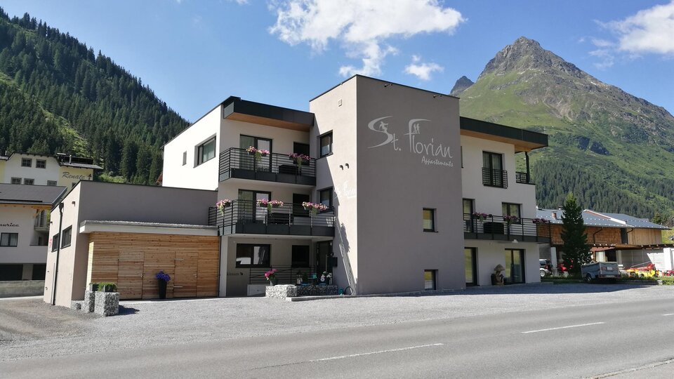 A modern building in the mountains with a clear blue sky. The house contains several apartments and is surrounded by green forests.