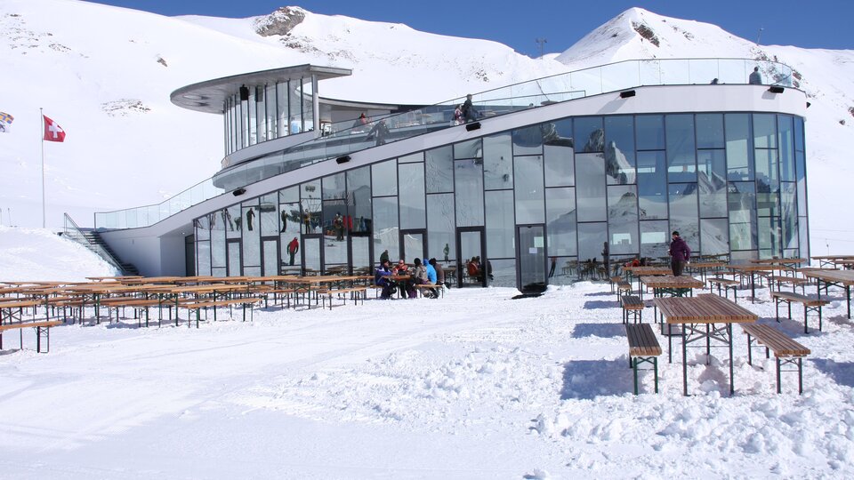 A modern building in the mountains, surrounded by snow. Outside, there are rows of wooden benches and people enjoying the wintry landscape.