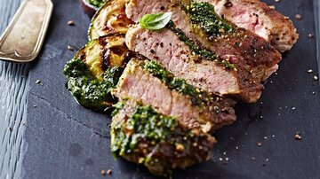 A delicious piece of meat, perfectly cooked and garnished with green pesto. Grilled zucchini are served on a dark serving board.