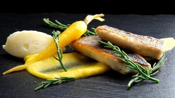An artfully arranged plate with grilled fish, surrounded by colorful puree and fresh vegetables. The presentation uses elegant elements such as herbs and a dark base for a modern look.