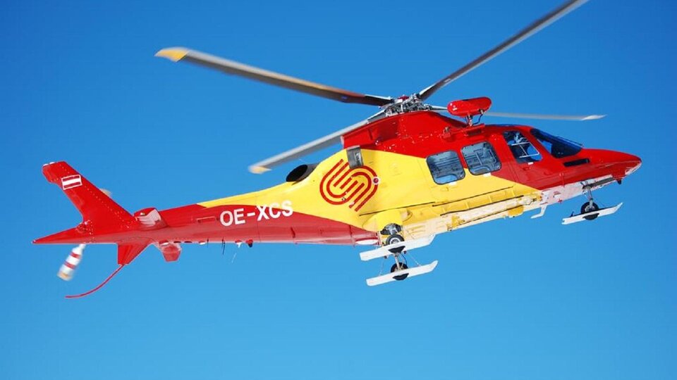 A colorful helicopter flies against a clear blue sky. The landing gear is clearly visible.