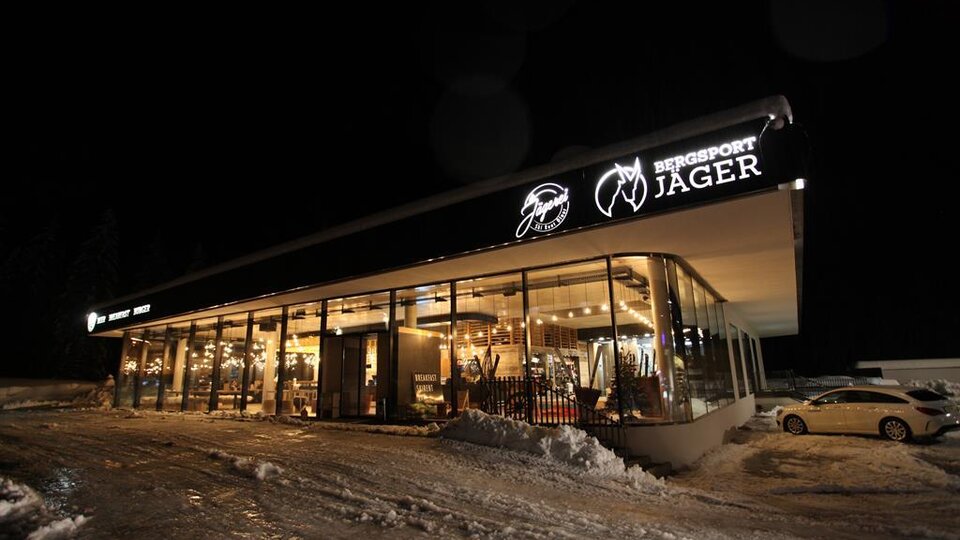 A modern sports shop with large windows, well-lit at night. It is surrounded by snow and the store name "Bergsport Jager" is clearly visible.