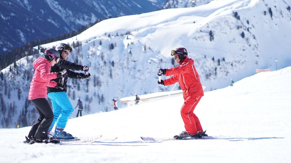 A ski school in the mountains with three people learning to ski. The surroundings are covered in snow and the mountains are impressive.