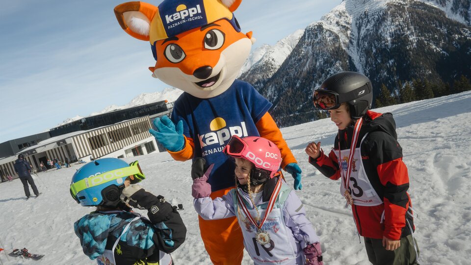 A mascot in a blue shirt and a hat stands on a snow-covered slope. Around it, three children in ski gear are celebrating together.
