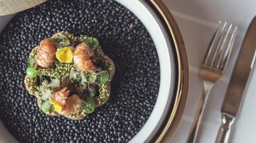 An artfully arranged dish with shrimp, herbs, and colorful ingredients on a black base. It is elegantly presented and looks very appealing.