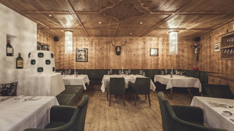Wood-paneled dining area with set tables, green upholstered chairs, warm lighting, and decorative elements such as deer antlers and wall paintings. | © Hannes Niederkofler