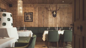 Wood-paneled dining area with set tables, green upholstered chairs, tiled stove, deer antlers, and warm light.