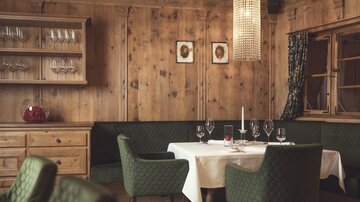Wood-paneled dining room with a laid table, green upholstered chairs, a glass cabinet, wall pictures, and warm lighting. | © Hannes Niederkofler