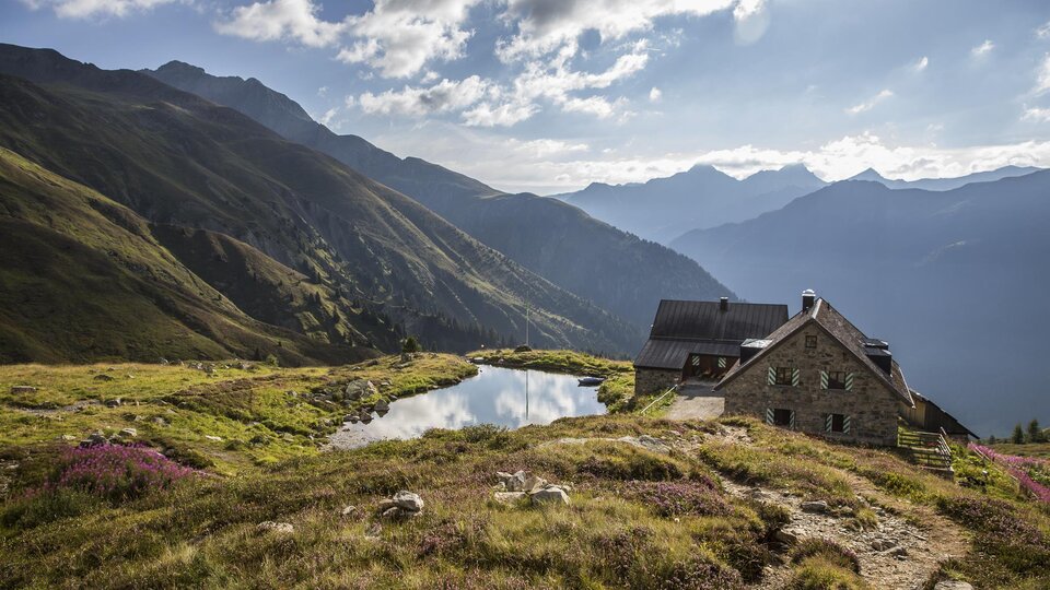 A picturesque mountain house overlooking green meadows and tall mountains. A tranquil mountain lake reflects the impressive scenery.
