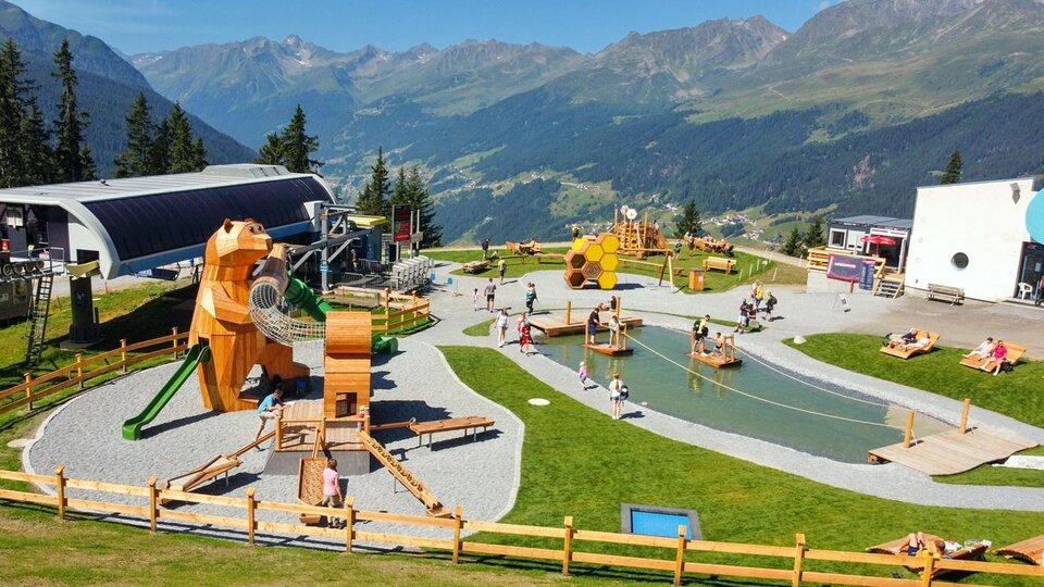 A playground in the mountains with various wooden play equipment for children. In the background, green meadows and tall mountains can be seen.