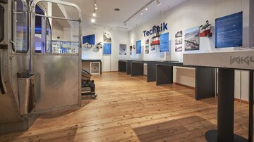 A modern exhibition space with wooden flooring and blue information walls. Various exhibits and tables are arranged in the room.