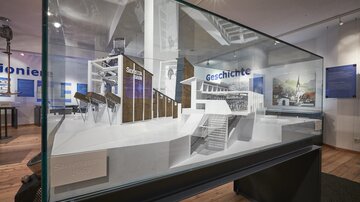 A display case with an architectural model showcasing various buildings. The background contains informative texts and images about the history.