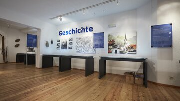A modern room with wooden flooring that showcases an exhibition on the topic of history. Explanatory panels and pictures are hanging on the wall.