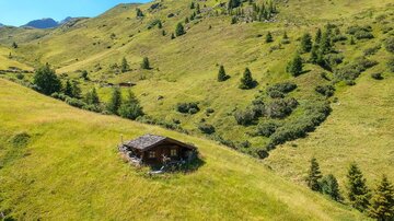 A picturesque cabin stands on a gentle meadow in the mountains. Green hills and clear blue skies surround the scene.