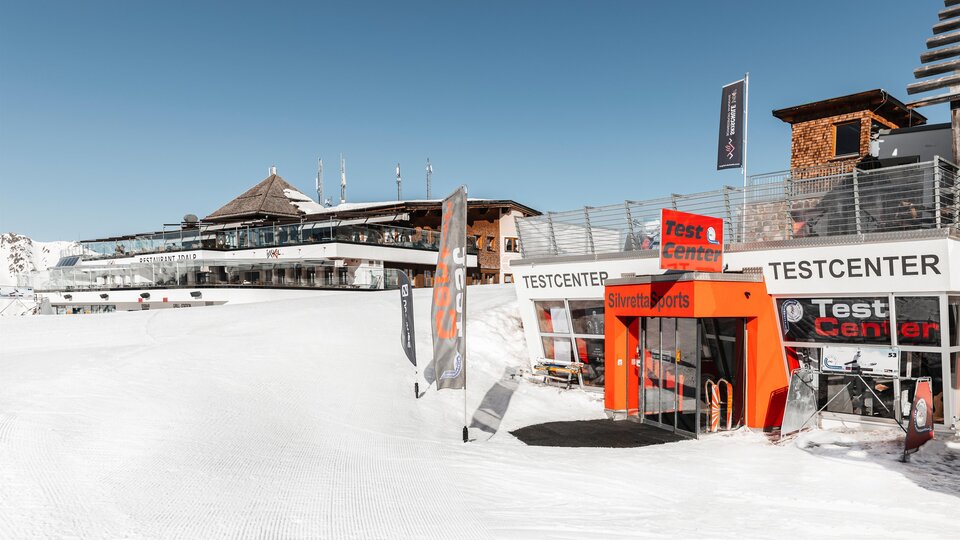 Ein schneebedecktes Skigebiet mit einem Testzentrum und einem modernen Gebäude im Hintergrund. Der Himmel ist klar und blau, ideal für Wintersportaktivitäten.