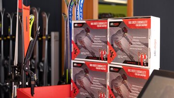 Eine Präsentation von Skiausrüstung in einem Geschäft. Boxen mit Werbung für Produkte stehen auf einem roten Tisch.