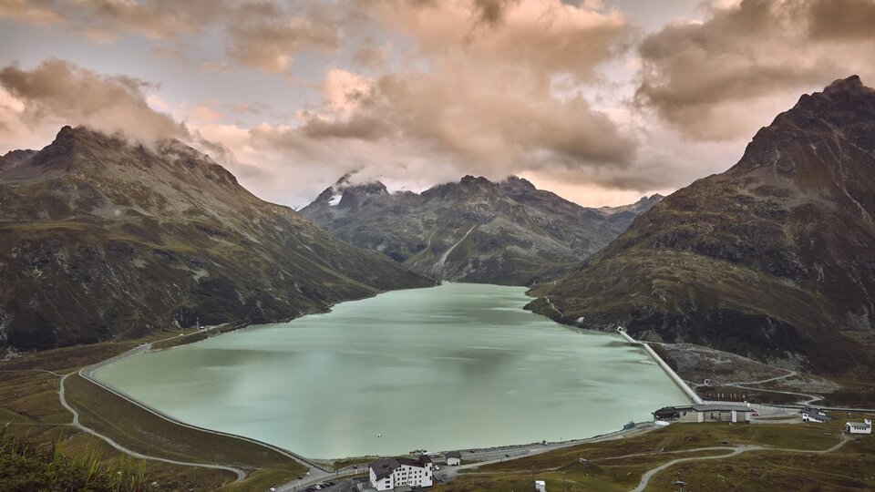 A beautiful mountain lake, surrounded by tall mountains and dramatic clouds. The calm water surface reflects the surrounding landscape.