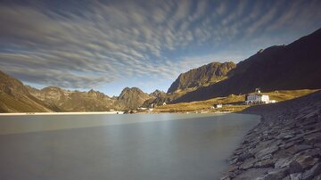 A tranquil lakescape surrounded by high mountains and dramatic cloud formations. In the foreground, a rocky shoreline with a white building is visible.