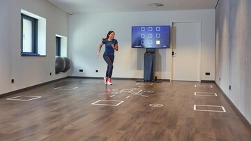 A woman is training in a modern room with a wooden floor. There is a screen on the wall, and on the floor, there are various marked areas.