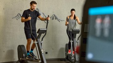 Two people are training in a modern gym on cross-trainers. The room has a plain wall and offers a motivating atmosphere.